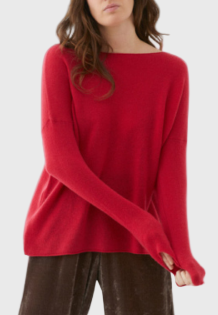 A woman standing in a studio setting, wearing a slightly oversized red cashmere sweater paired with brown trousers.
