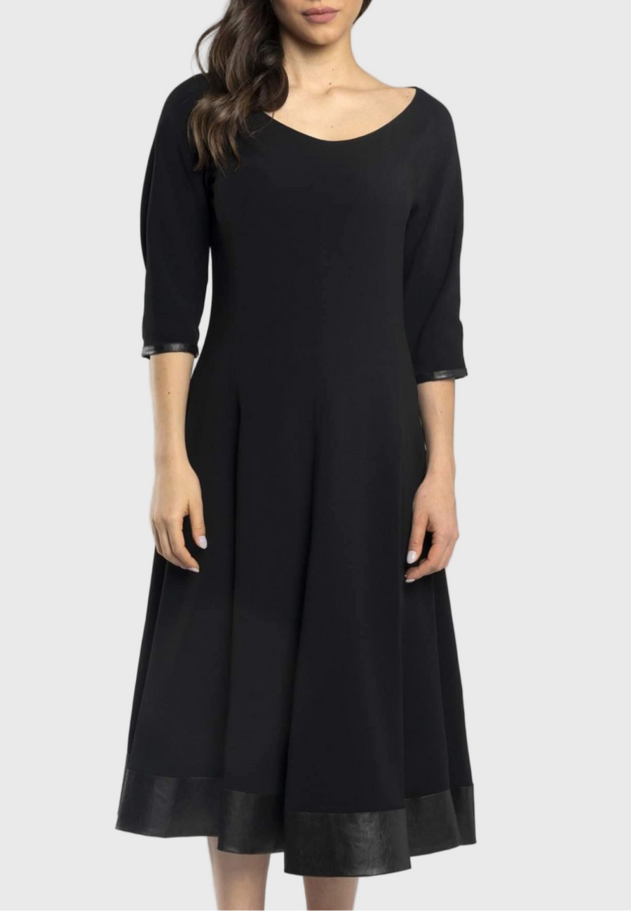 A woman wearing a black midi dress with a round neckline, mid-length sleeves, and a contrasting black eco-leather panel at the hem.